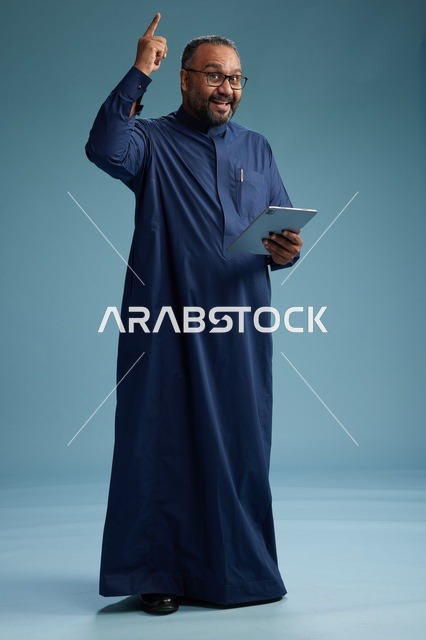 بورتريه لرجل عربي سعودي  خليجي بثوب أزرق يستخدم جهاز لوحي, استخدام التابليت المحمول, العمل اونلاين, الجسم بالكامل, خلفيه زرقاء