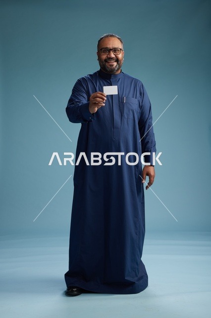 بورتريه لرجل عربي  سعودي  خليجي يرتدي ثوباً أزرق يمسك بيده بطاقة بيضاء فارغه, الجسم بالكامل, خلفيه زرقاء
