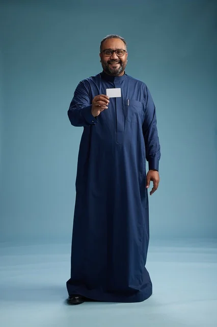 A portrait of an Arab man, Saudi, Gulf, wearing a blue thobe, holding a blank white card in his hand, full body, blue background.