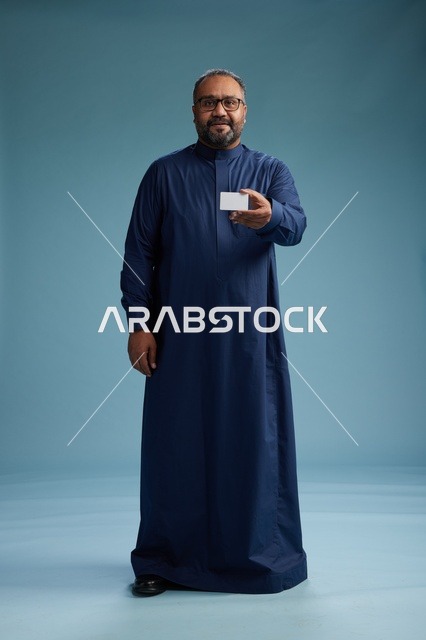 بورتريه لرجل عربي  سعودي  خليجي يرتدي ثوباً أزرق يمسك بيده بطاقة بيضاء فارغه, الجسم بالكامل, خلفيه زرقاء