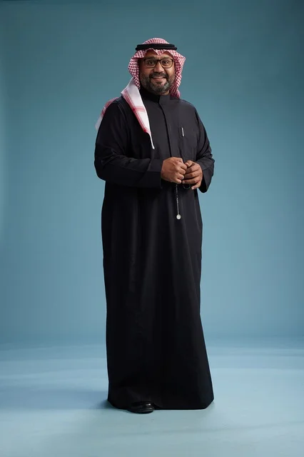 A smiling Saudi man wearing a black thobe with a shemagh and agal, standing confidently with his full body, blue background.