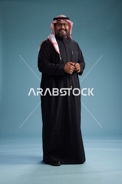 رجل سعودي مبتسم يرتدي ثوب أسود مع شماغ وعقال, الوقوف بثقه لكامل الجسم, خلفيه زرقاء