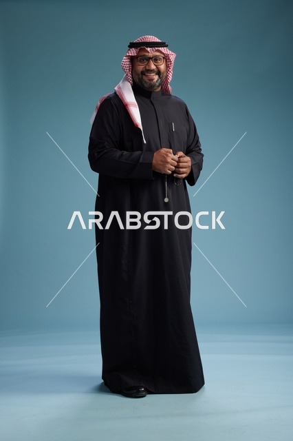 رجل سعودي مبتسم يرتدي ثوب أسود مع شماغ وعقال, الوقوف بثقه لكامل الجسم, خلفيه زرقاء