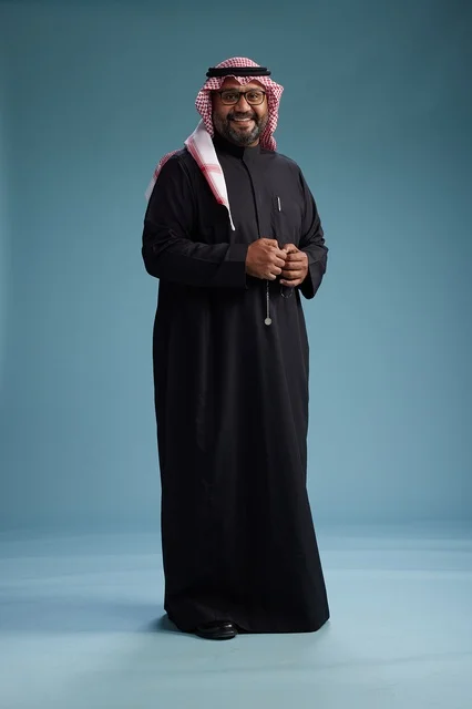 A smiling Saudi man wearing a black thobe with a shemagh and agal, standing confidently with his full body, blue background.