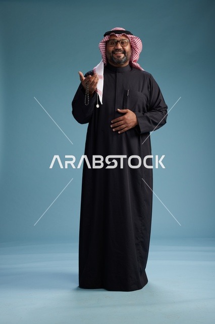 رجل سعودي مبتسم يرتدي ثوب أسود مع شماغ وعقال, الوقوف بثقه لكامل الجسم, خلفيه زرقاء