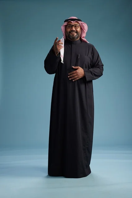A smiling Saudi man wearing a black thobe with a shemagh and agal, standing confidently with his full body, blue background.