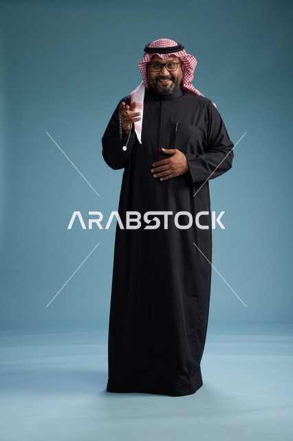 رجل سعودي مبتسم يرتدي ثوب أسود مع شماغ وعقال, الوقوف بثقه لكامل الجسم, خلفيه زرقاء