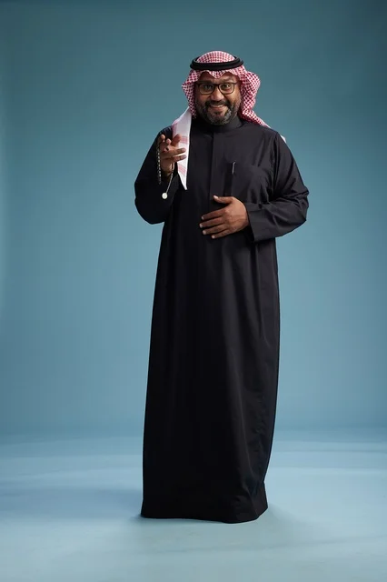 A smiling Saudi man wearing a black thobe with a shemagh and agal, standing confidently with his full body, blue background.
