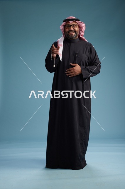 رجل سعودي مبتسم يرتدي ثوب أسود مع شماغ وعقال, الوقوف بثقه لكامل الجسم, خلفيه زرقاء