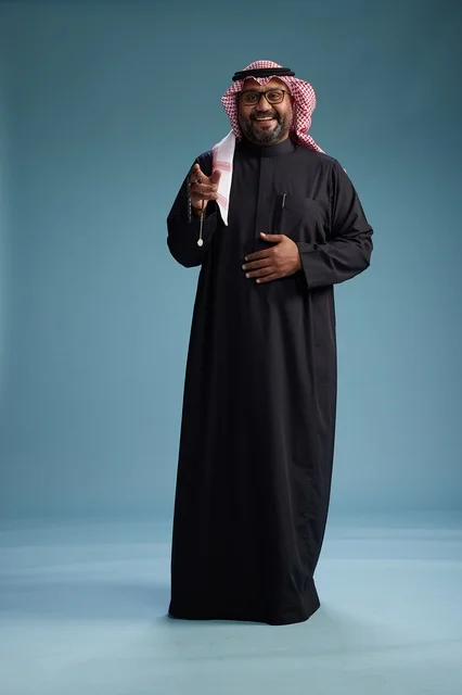 A smiling Saudi man wearing a black thobe with a shemagh and agal, standing confidently with his full body, blue background.