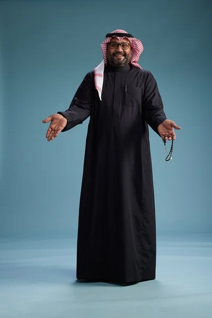 A smiling Saudi man wearing a black thobe with a shemagh and agal, standing confidently with his full body, blue background.