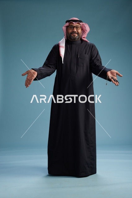 رجل سعودي مبتسم يرتدي ثوب أسود مع شماغ وعقال, الوقوف بثقه لكامل الجسم, خلفيه زرقاء