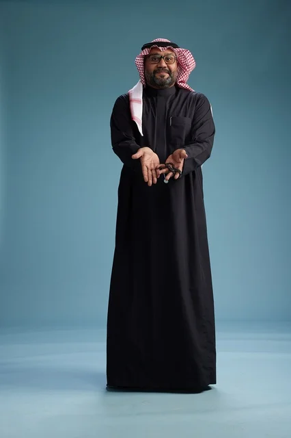 A smiling Saudi man wearing a black thobe with a shemagh and agal, standing confidently with his full body, blue background.