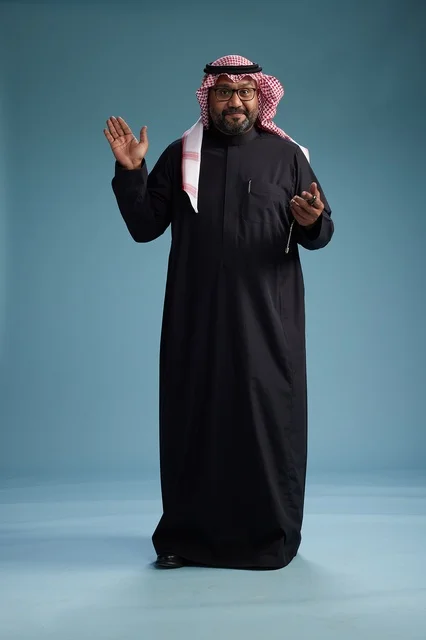 A smiling Saudi man wearing a black thobe with a shemagh and agal, standing confidently with his full body, blue background.