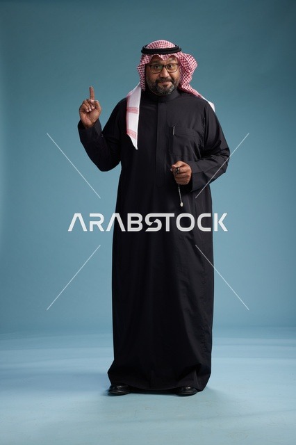 رجل سعودي مبتسم يرتدي ثوب أسود مع شماغ وعقال, الوقوف بثقه لكامل الجسم, خلفيه زرقاء