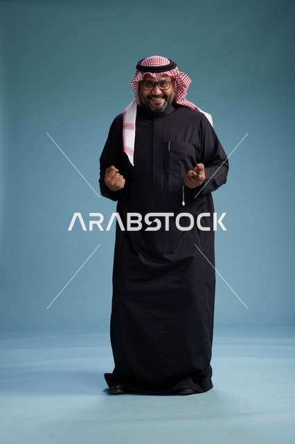 رجل سعودي مبتسم يرتدي ثوب أسود مع شماغ وعقال, الوقوف بثقه لكامل الجسم, خلفيه زرقاء