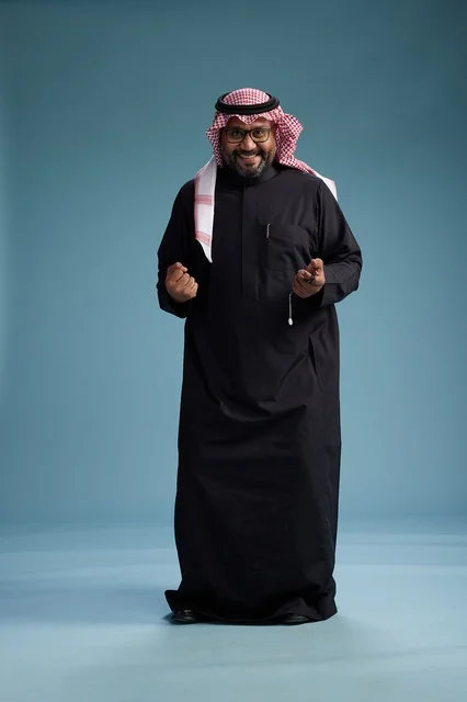 A smiling Saudi man wearing a black thobe with a shemagh and agal, standing confidently with his full body, blue background.