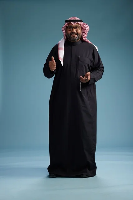 A smiling Saudi man wearing a black thobe with a shemagh and agal, standing confidently with his full body, blue background.