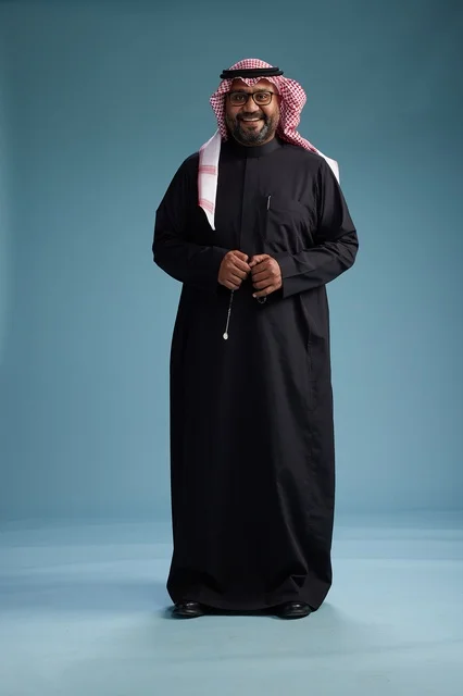 A smiling Saudi man wearing a black thobe with a shemagh and agal, standing confidently with his full body, blue background.