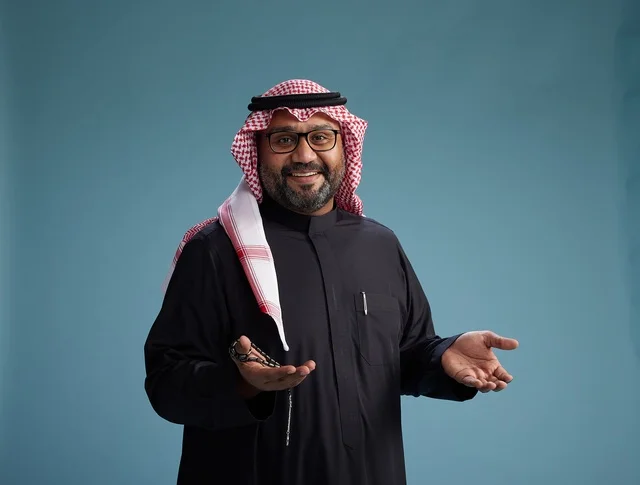 A smiling Saudi man wearing a black thobe with a shemagh and agal, with various hand gestures and movements, against a blue background.