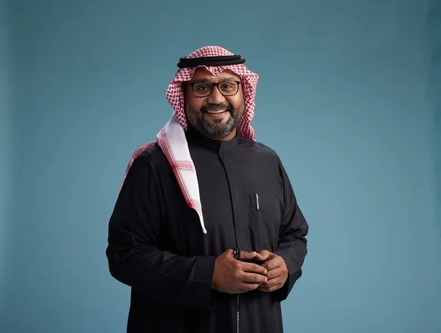 A smiling Saudi man wearing a black thobe with a shemagh and agal, with various hand gestures and movements, against a blue background.