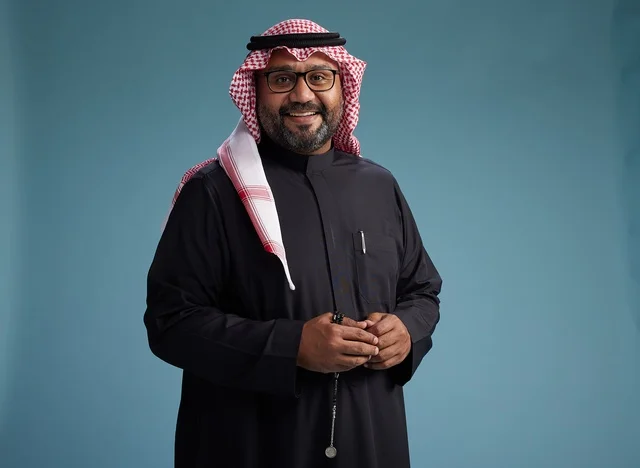A smiling Saudi man wearing a black thobe with a shemagh and agal, with various hand gestures and movements, against a blue background.