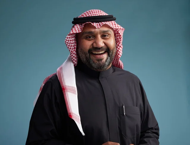 A smiling Saudi man wearing a black thobe with a shemagh and agal, with various hand gestures and movements, against a blue background.