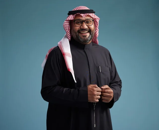 A smiling Saudi man wearing a black thobe with a shemagh and agal, with various hand gestures and movements, against a blue background.