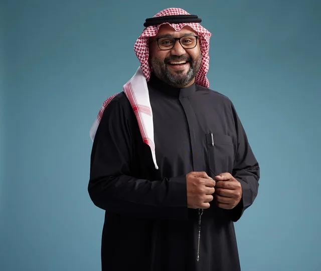 A smiling Saudi man wearing a black thobe with a shemagh and agal, with various hand gestures and movements, against a blue background.