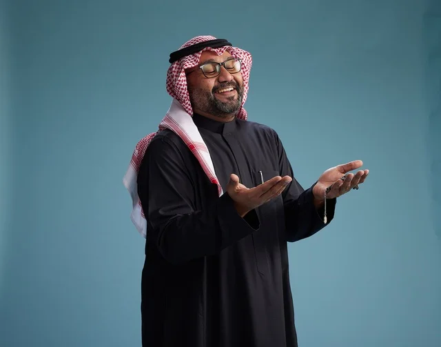 A smiling Saudi man wearing a black thobe with a shemagh and agal, with various hand gestures and movements, against a blue background.