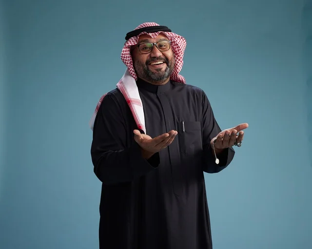 A smiling Saudi man wearing a black thobe with a shemagh and agal, with various hand gestures and movements, against a blue background.