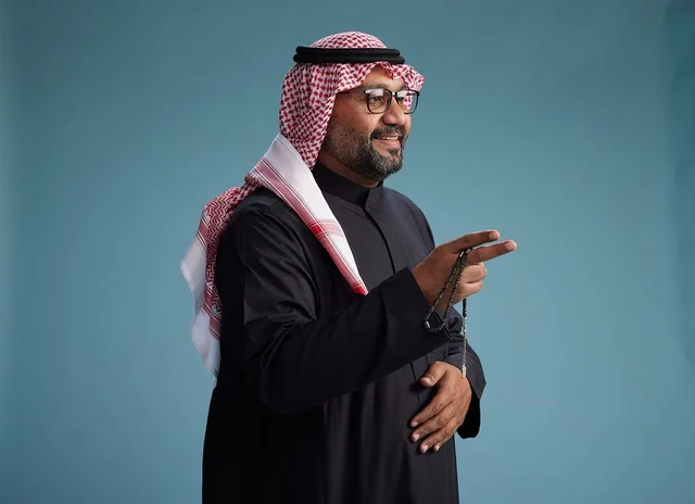 A smiling Saudi man wearing a black thobe with a shemagh and agal, with various hand gestures and movements, against a blue background.