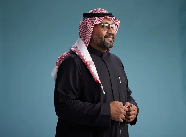 A smiling Saudi man wearing a black thobe with a shemagh and agal, with various hand gestures and movements, against a blue background.