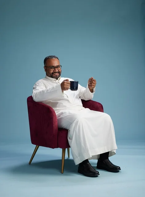 A portrait of a Saudi man laughing and enjoying coffee while sitting, with gestures of happiness and joy, against a blue background.