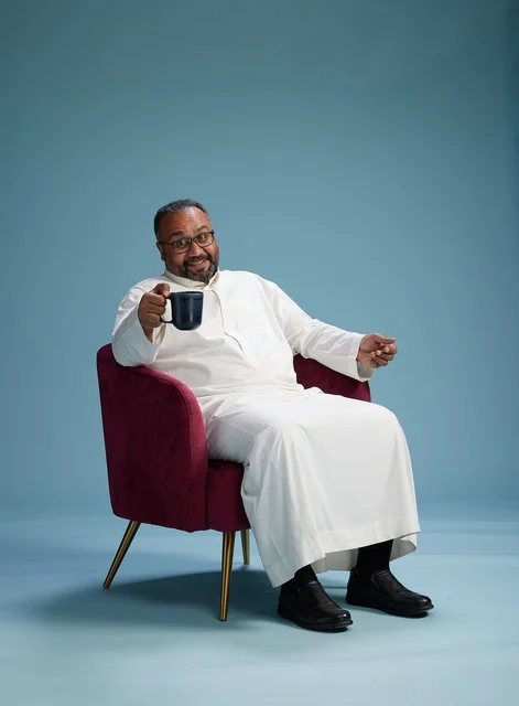 A portrait of a Saudi man laughing and enjoying coffee while sitting, with gestures of happiness and joy, against a blue background.