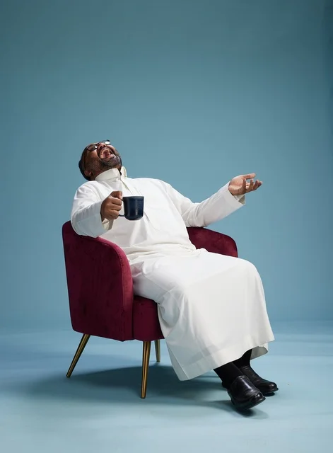 بورتريه لرجل سعودي يضحك ويستمتع بالقهوة وهو جالس, ايماءات السعاده والسرور, خلفيه زرقاء