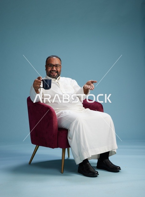 A portrait of a Saudi man laughing and enjoying coffee while sitting, with gestures of happiness and joy, against a blue background.