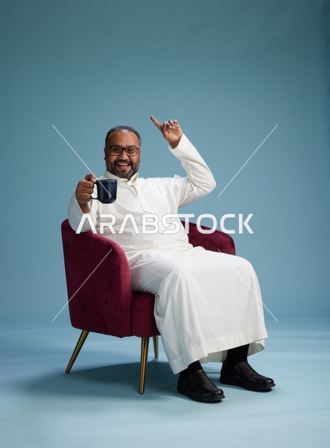 A portrait of a Saudi man laughing and enjoying coffee while sitting, with gestures of happiness and joy, against a blue background.