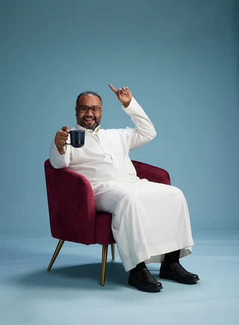 A portrait of a Saudi man laughing and enjoying coffee while sitting, with gestures of happiness and joy, against a blue background.