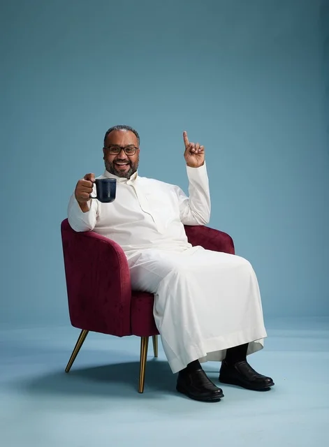 A portrait of a Saudi man laughing and enjoying coffee while sitting, with gestures of happiness and joy, against a blue background.