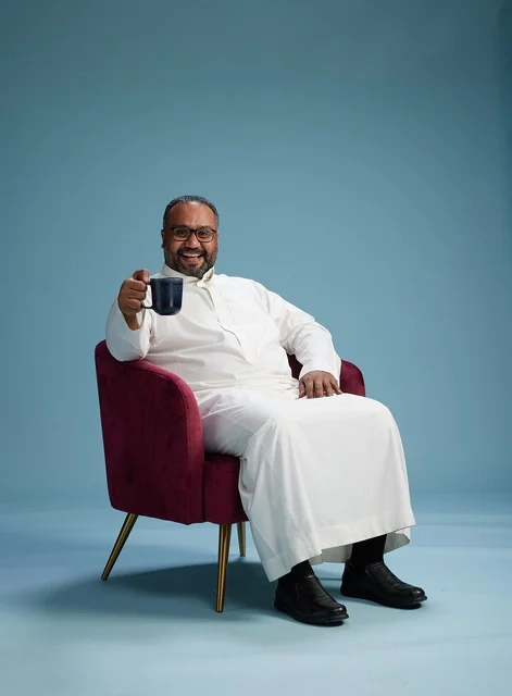 بورتريه لرجل سعودي يضحك ويستمتع بالقهوة وهو جالس, ايماءات السعاده والسرور, خلفيه زرقاء