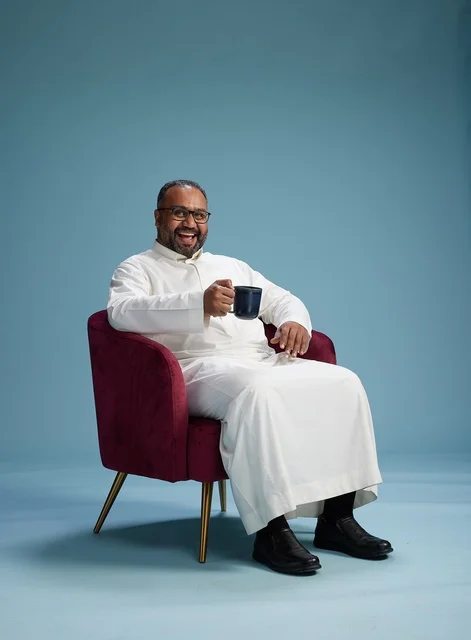 A portrait of a Saudi man laughing and enjoying coffee while sitting, with gestures of happiness and joy, against a blue background.