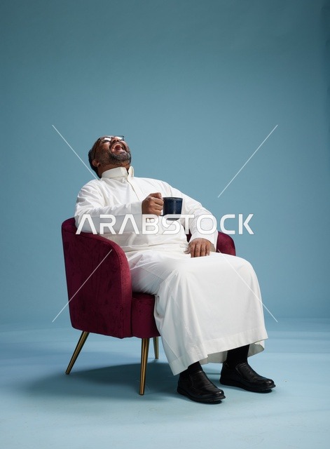 بورتريه لرجل سعودي يضحك ويستمتع بالقهوة وهو جالس, ايماءات السعاده والسرور, خلفيه زرقاء