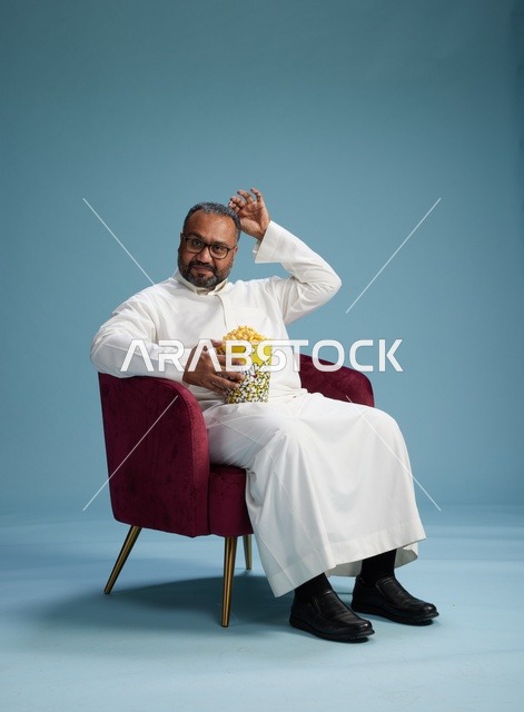 بورتريه لرجل  عربي سعودي خليجي يرتدي الثوب الابيض  يجلس على كرسي مريح, الاستمتاع بتناول  الفشار , خلفيه زرقاء