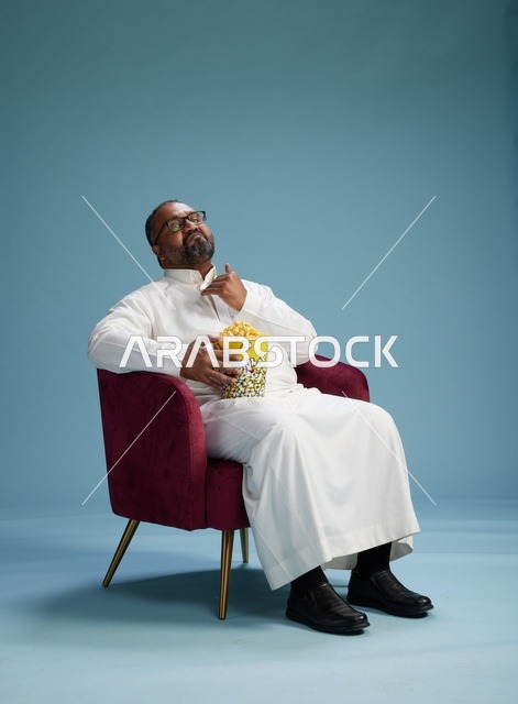 بورتريه لرجل  عربي سعودي خليجي يرتدي الثوب الابيض  يجلس على كرسي مريح, الاستمتاع بتناول  الفشار , خلفيه زرقاء