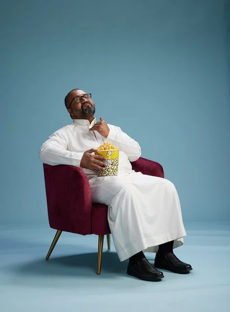 A portrait of an Arab Saudi Gulf man wearing a white thobe sitting on a comfortable chair, enjoying eating popcorn, with a blue background.