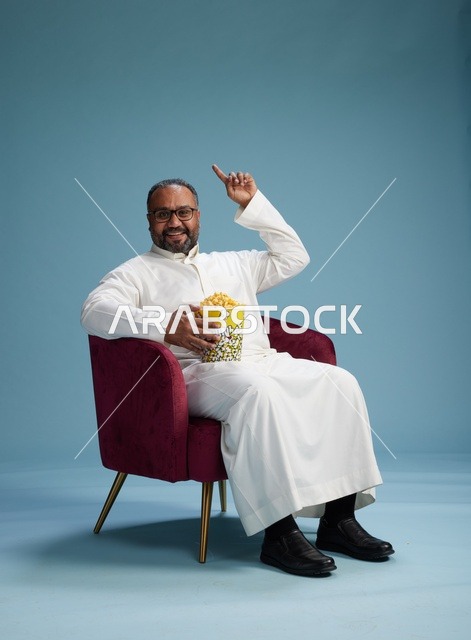 A portrait of an Arab Saudi Gulf man wearing a white thobe sitting on a comfortable chair, enjoying eating popcorn, with a blue background.
