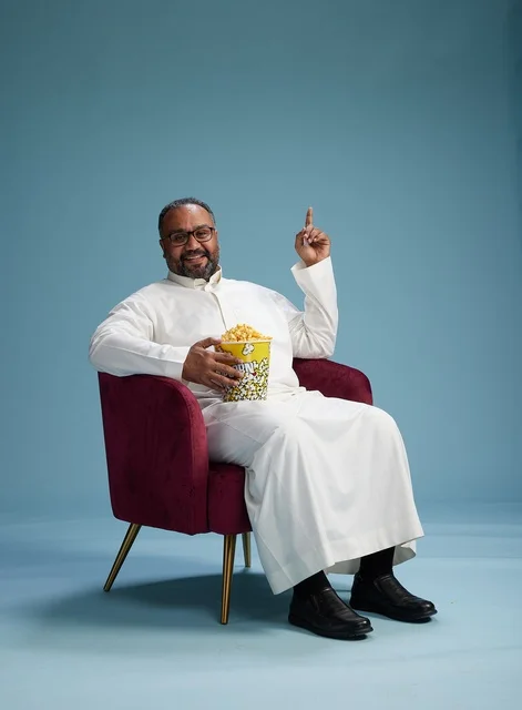 A portrait of an Arab Saudi Gulf man wearing a white thobe sitting on a comfortable chair, enjoying eating popcorn, with a blue background.