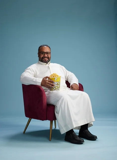 A portrait of an Arab Saudi Gulf man wearing a white thobe sitting on a comfortable chair, enjoying eating popcorn, with a blue background.