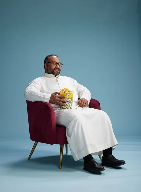 A portrait of an Arab Saudi Gulf man wearing a white thobe sitting on a comfortable chair, enjoying eating popcorn, with a blue background.
